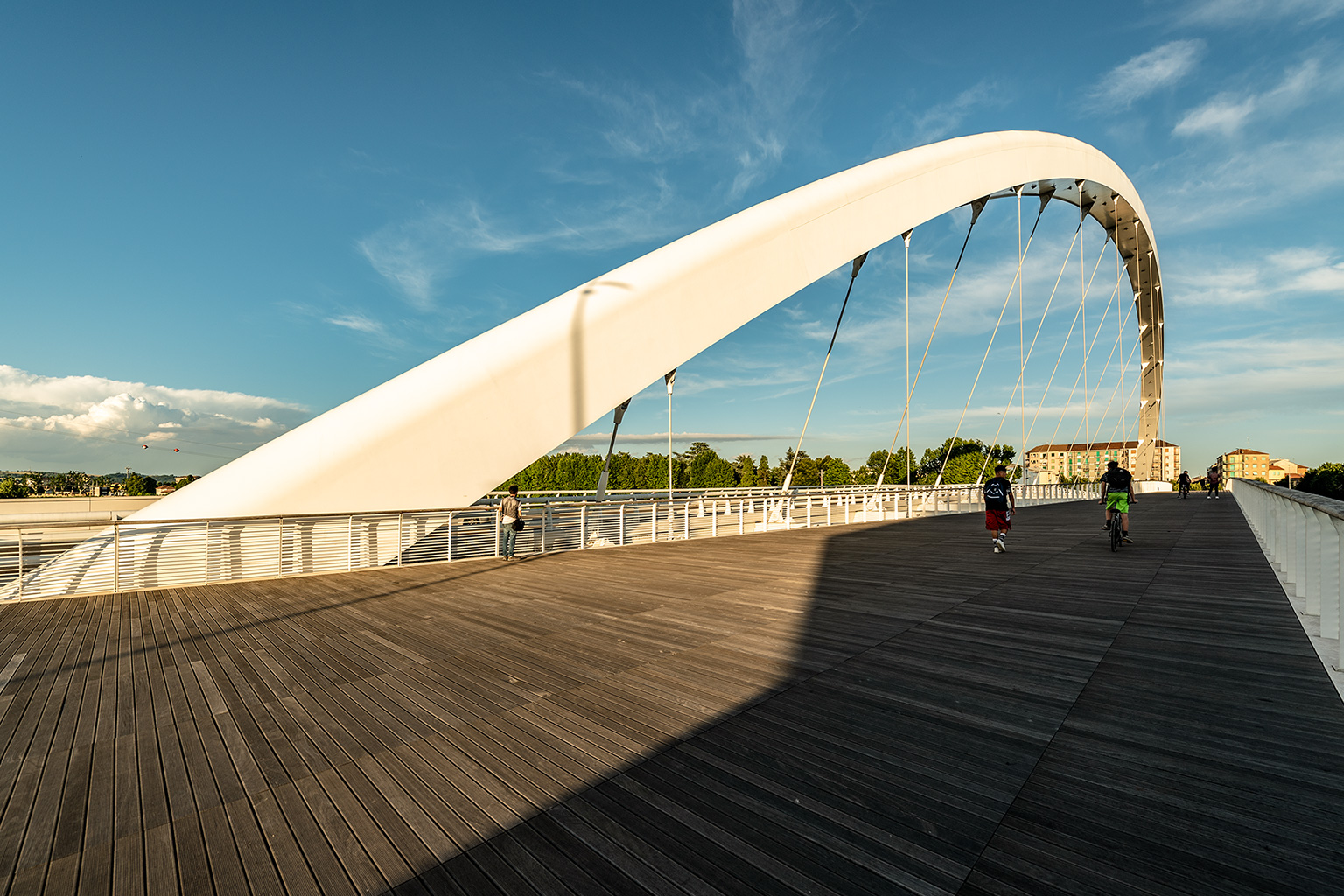 Cittadella Bridge, Alessandria. Perspective view of the Cittadella Bridge in Alessandria during the golden afternoon hours, featuring the iconic inclined white arch, steel tension cables, and a wooden plank walkway, with pedestrians and cyclists crossing.