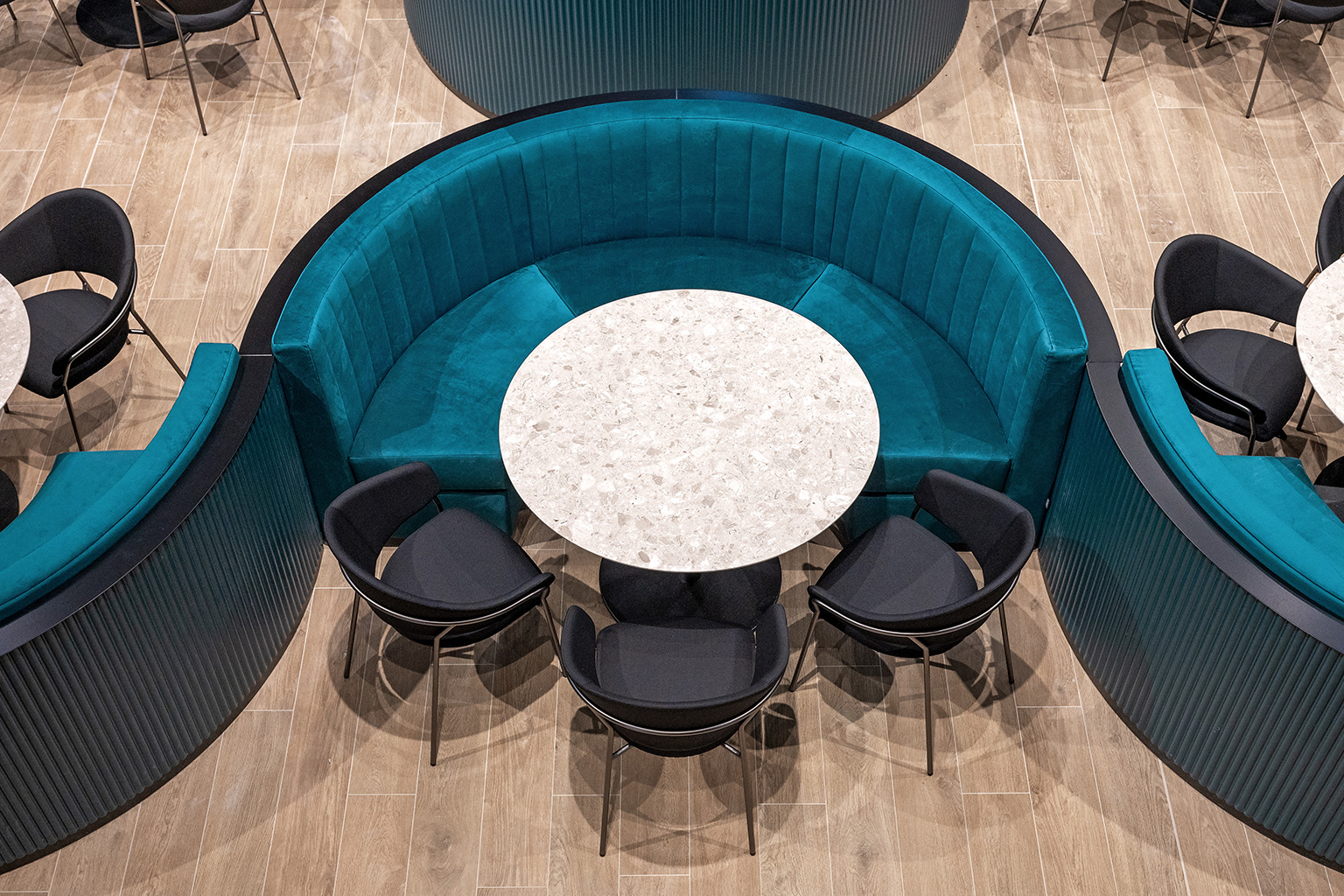 Ristopub ChorusLife, Bergamo. Top view of a modern lounge area featuring a semicircular turquoise velvet bench, a round table with a light stone top, and black chairs on a light wood floor.