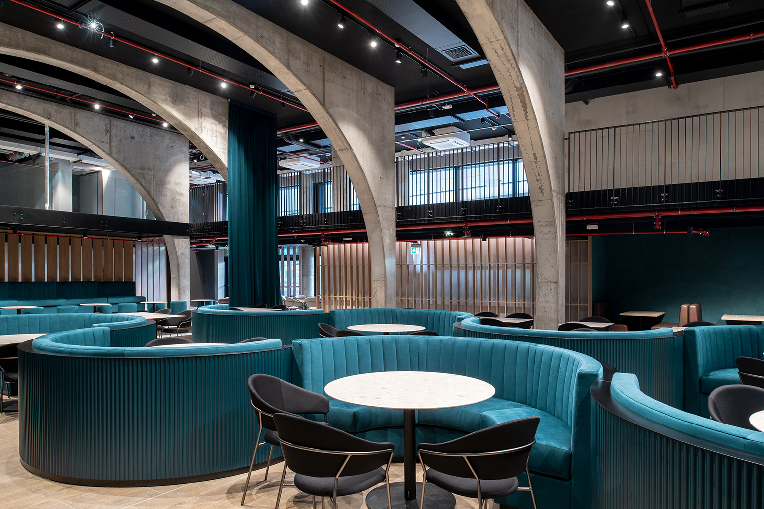 Ristopub ChorusLife, Bergamo. Interior of a modern restaurant with semicircular turquoise velvet seating, round tables with light stone tops, black chairs, and large exposed concrete arches; the space is open and airy, featuring a dark ceiling, exposed red piping, and technical lighting.