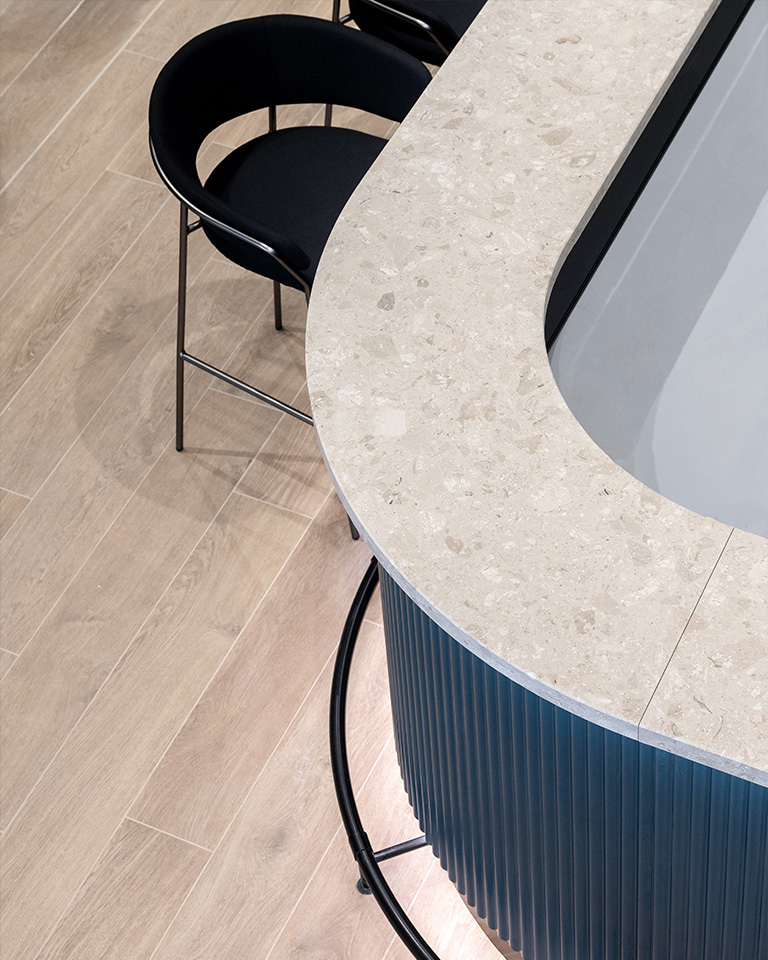 Ristopub ChorusLife, Bergamo. Close-up view of a modern curved bar counter with a light stone surface and ribbed blue base, accompanied by black upholstered bar stools on a light wood floor.