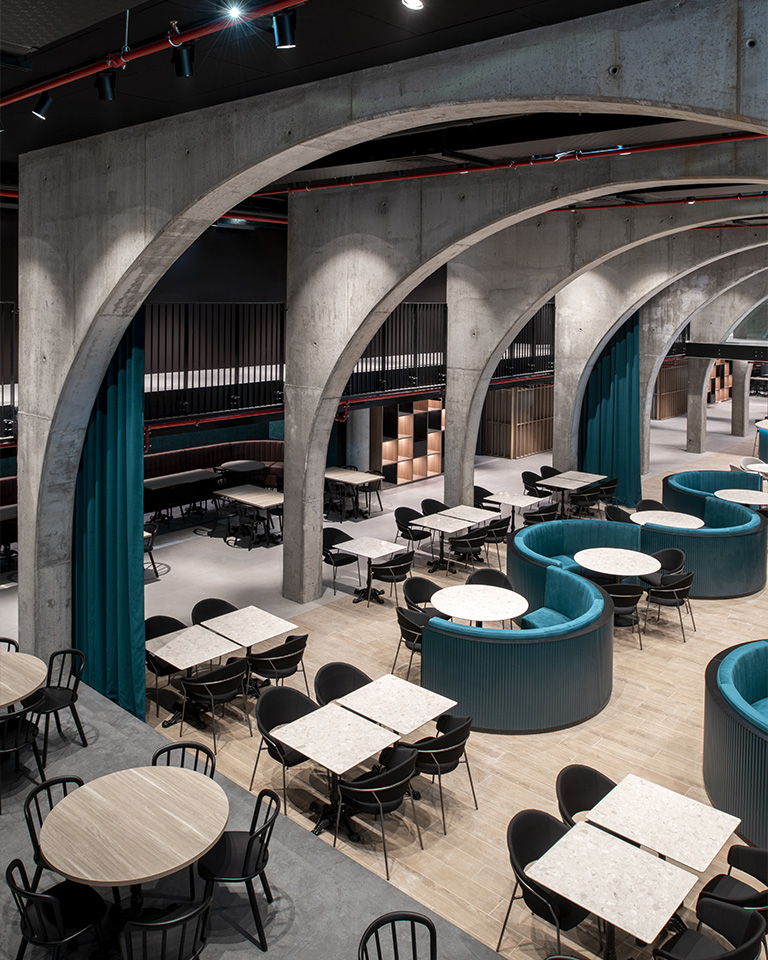 Ristopub ChorusLife, Bergamo. Panoramic view of the interior featuring exposed concrete arches, light marble tables with black chairs, and curved upholstered seating in turquoise velvet; the space blends industrial elements with elegant furnishings in a spacious and refined environment.