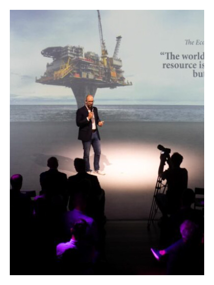 Circolo delle Imprese, Milan. Relatore su un palco illuminato presenta davanti a un pubblico seduto, con la proiezione di una piattaforma petrolifera offshore sullo sfondo e una citazione sul valore della conoscenza come risorsa strategica.